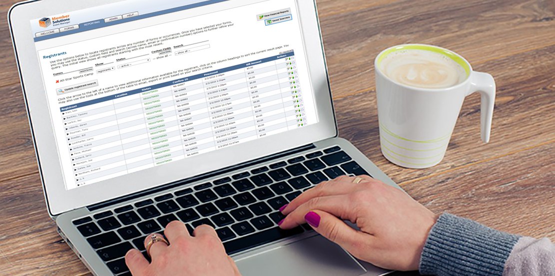 Woman using saved searches feature of Event Manager software to track registrations on laptop. Close view over shoulder of wooden desk with coffee.